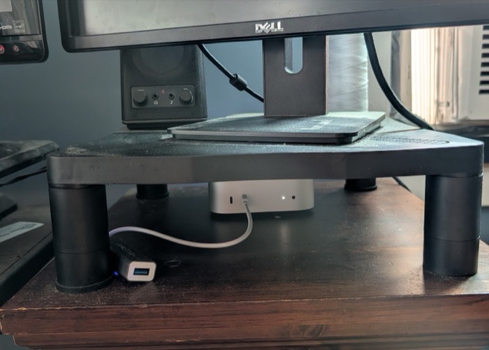 A Mac Mini on a wooden desk beneath a monitor stand, a Dell monitor above it, a single white cable running to a small USB hub.